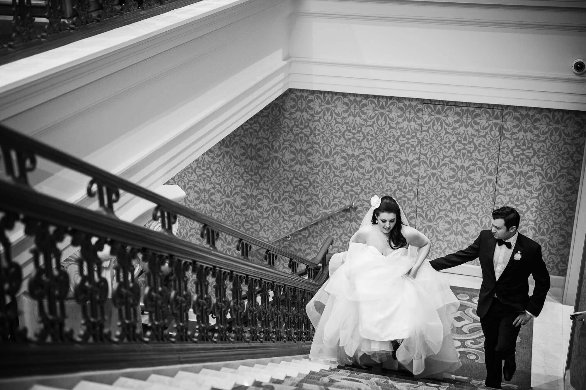 Black and white portrait of the groom helping the bride carry her dress up the stairs at the Omni King Edward Hotel in Toronto