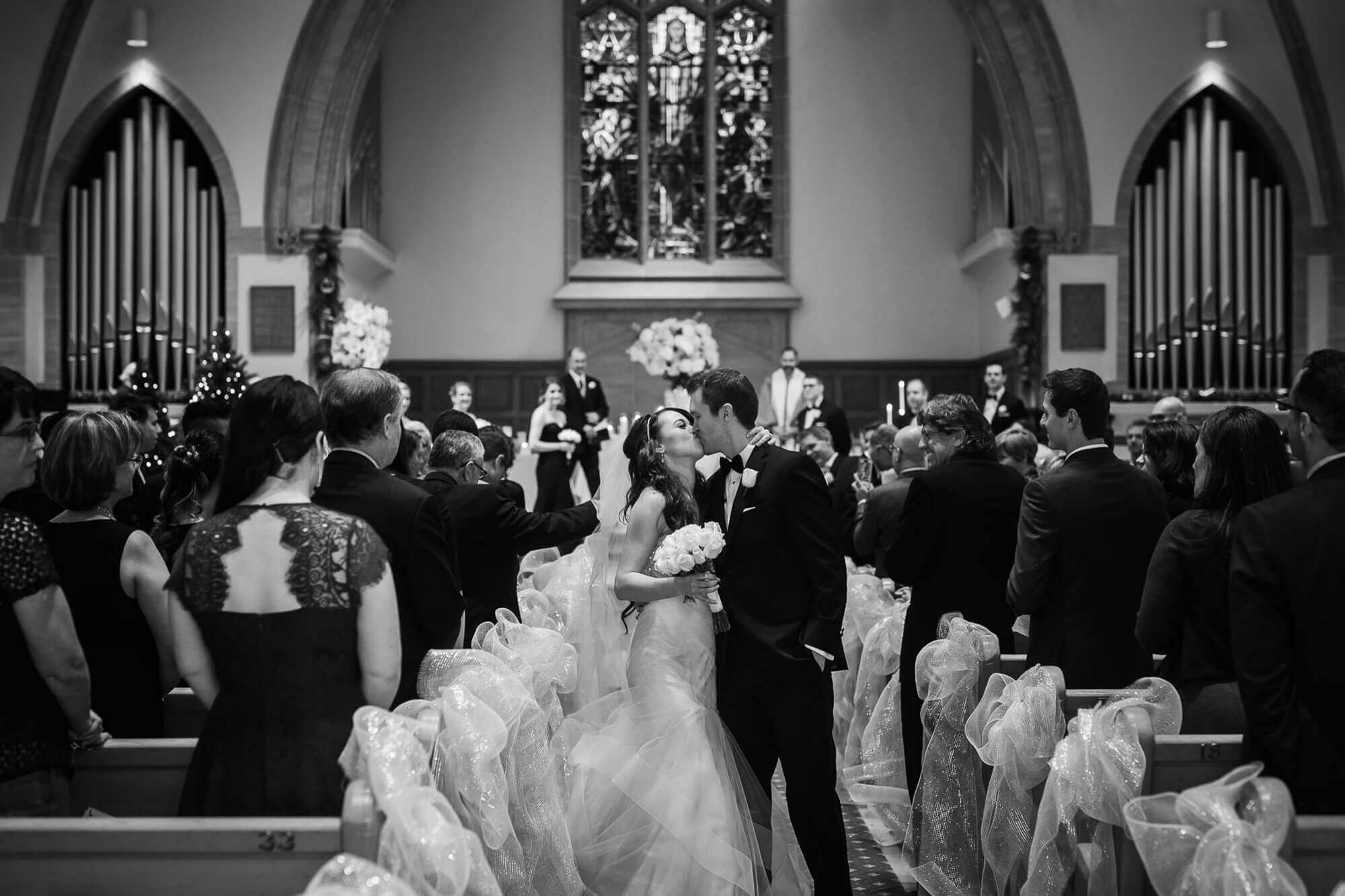 Black and white portrait of the bride and groom kissing down the aisle of the Rosedale United Church in Toronto
