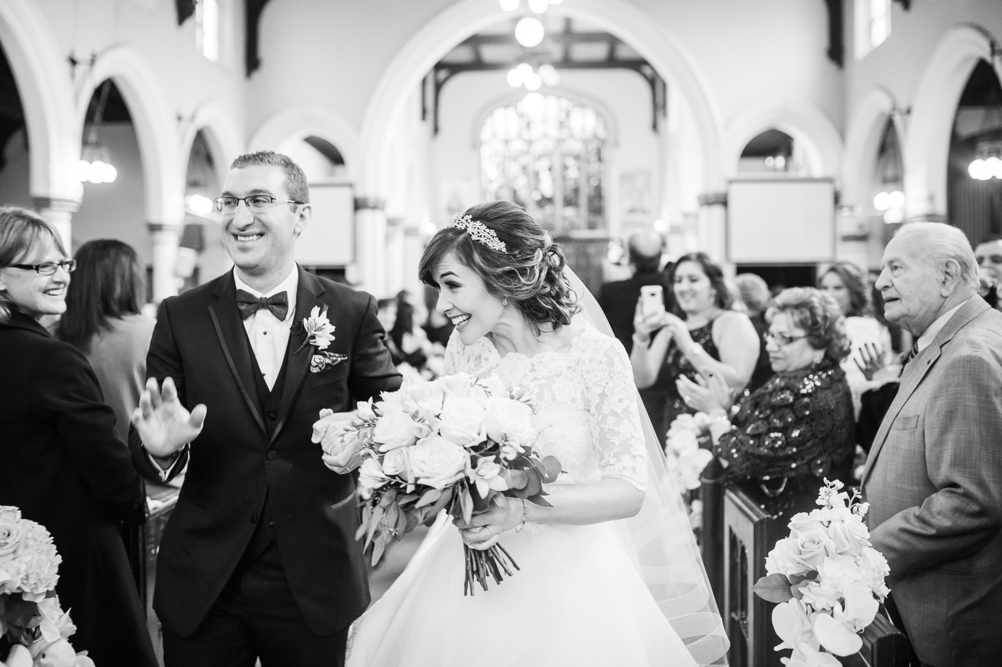 Black and white portrait of the bride and groom waving to their guests as they exit Our Lady of Lebanon